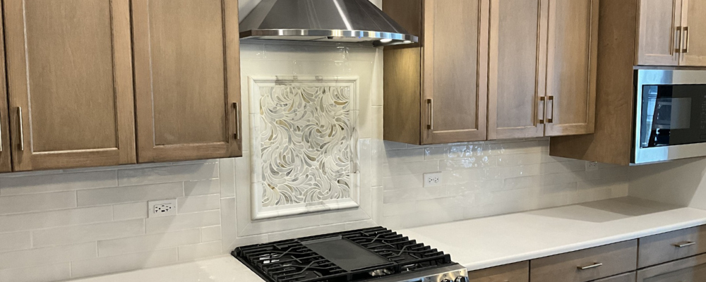 Modern kitchen with light wood cabinets, white tile backsplash, a black gas cooktop in the center, stainless range hood above, and a built-in microwave on the right.
